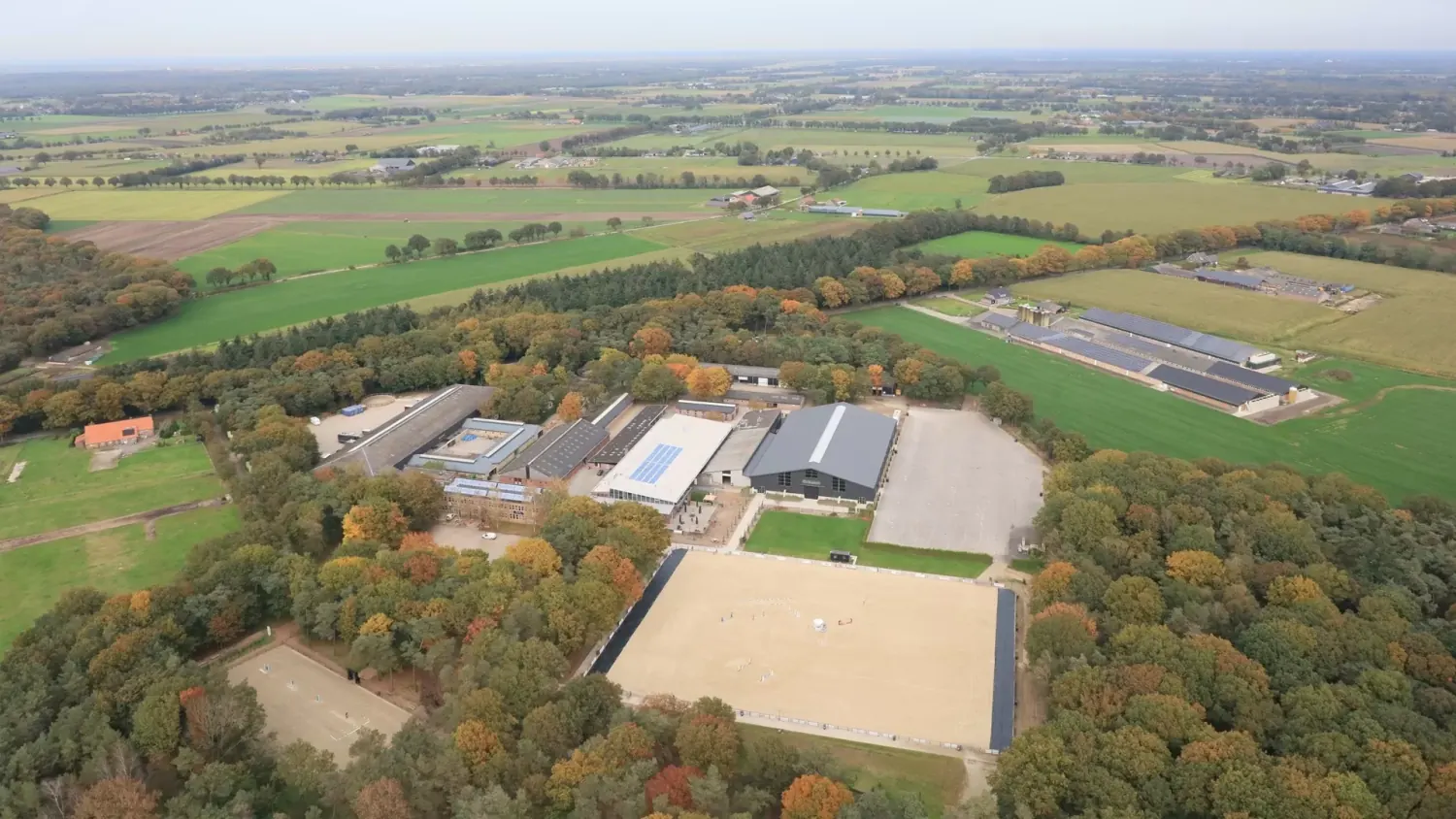 Luchtfoto van paardensportcentrum Green Valley Estate in Deurne, met de grote zandbak, binnenmanege en stallen tussen herfstbossen en weilanden