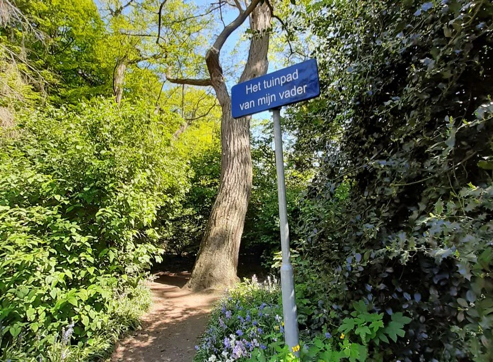 Bord “Het tuinpad van mijn vader” bij een zandpad tussen bomen en bloeiende boshyacinten