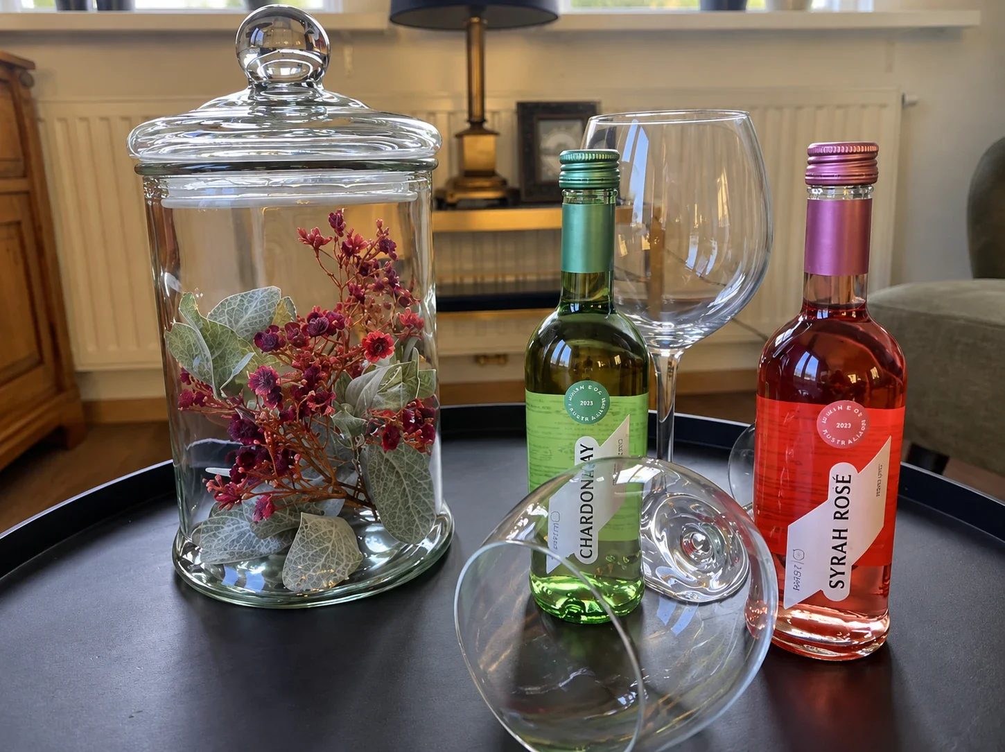 Welkomstdrankje op de salontafel: flesjes chardonnay en syrah rosé met wijnglazen naast een glazen pot met bloemen